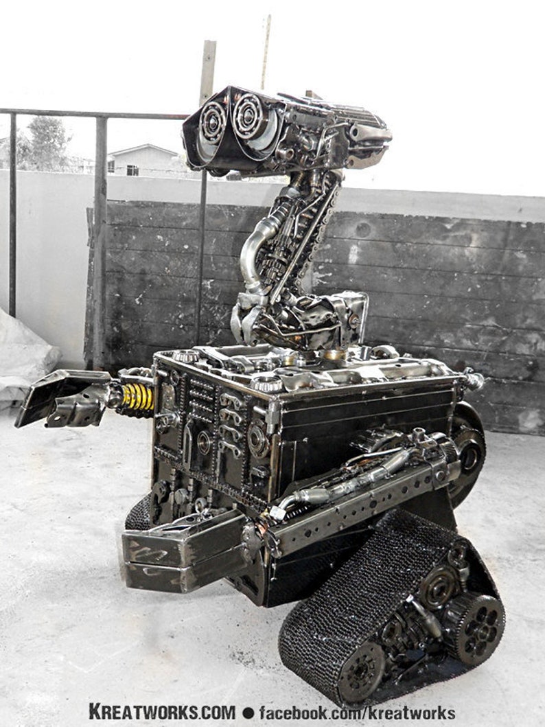 May include: A robot made entirely of recycled metal parts. The robot has a large head with two round eyes, a long neck, and a body with two tracks. The robot is made of various shades of gray metal.