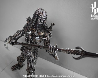 May include: A metal sculpture of a Predator, a fictional alien hunter, holding a spear. The sculpture is made of recycled metal parts and is highly detailed. The Predator is wearing a helmet and has a menacing expression.