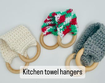 Puede incluir: Tres colgadores de toallas de cocina de ganchillo con aros de madera. Uno es blanquecino, otro rojo, verde y blanco, y el tercero es gris con hilo plateado. El texto "Kitchen towel hangers" está en la imagen.