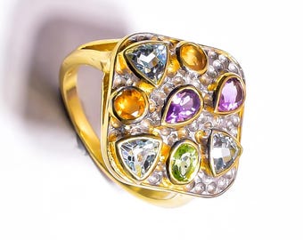 May include: A gold ring featuring a square-shaped face with a textured silver-toned surface. The ring is set with multiple multi-coloured gemstones, including light blue, yellow, purple, and green stones in various triangular and oval shapes.