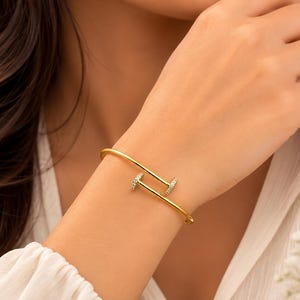 Può includere: Un bracciale rigido color oro a forma di chiodo, caratterizzato da una fascia liscia e curva che avvolge il polso. Le due estremità del design a forma di chiodo sono impreziosite da piccole pietre chiare e scintillanti sulle superfici piatte.