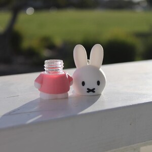 Pode incluir: Uma figura de coelho branco vestindo uma camisa rosa e contendo uma garrafa de vidro transparente. O coelho tem um nariz e uma boca pretos.