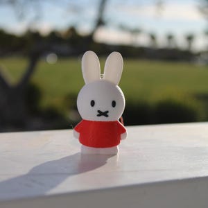Pode incluir: Uma figura de coelho branco vestindo uma camisa vermelha. O coelho tem olhos pretos e uma boca preta.