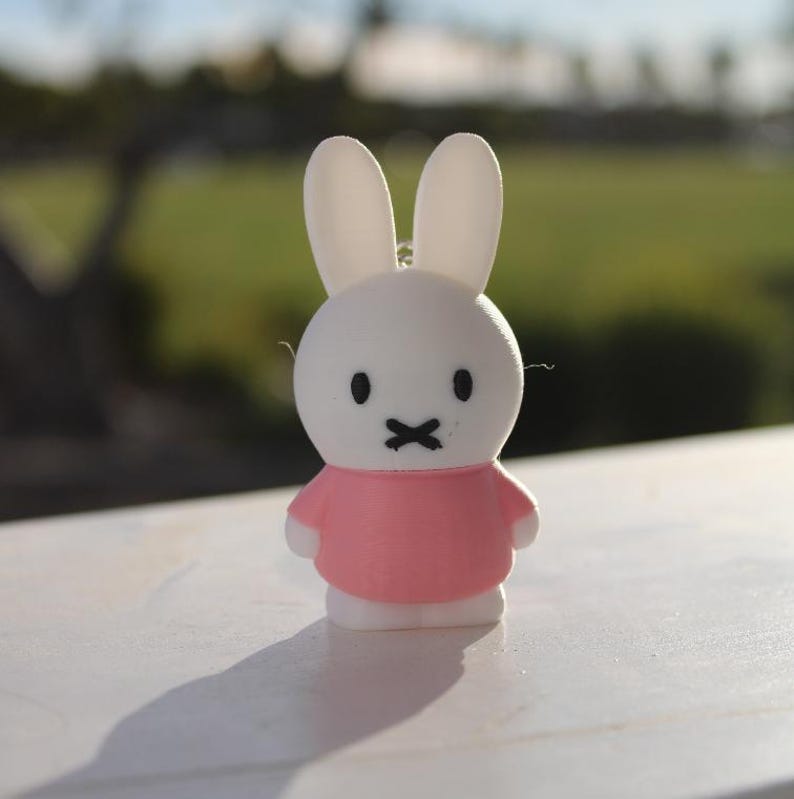 Pode incluir: Uma figura de coelho branco com roupa rosa e olhos pretos. O coelho tem uma pequena boca em forma de X preta.
