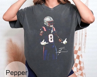 May include: Dark grey t-shirt featuring a football player in a helmet and uniform with the number 8. The player is in a dynamic pose with a signature below. The word "Pepper" is in a white circle at the bottom.