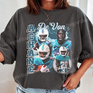May include: A charcoal gray short-sleeve t-shirt featuring a graphic print of football player De'Von Achane. The design includes multiple images of the player in a teal uniform, the text De'Von Achane, and a signature in a circular frame.