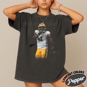 May include: Dark gray t-shirt featuring a football player in a green and yellow uniform. The image includes the text "NoRegretBae" and a signature. The shirt has a distressed look and the words "Comfort Colors Pepper" are in a circular design.