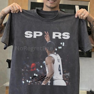 May include: Dark gray t-shirt featuring the word "SPARS" in white block letters. The shirt has a graphic of a basketball player making a peace sign. The player is wearing a white jersey with the number 4. The shirt also has the text "NoRegretBae".
