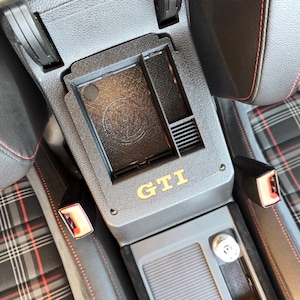 Könnte beinhalten: Schwarze Autokonsole mit den goldenen Buchstaben "GTI". Die Konsole hat ein eingebautes Staufach mit dem Volkswagen-Logo. Der Innenraum hat rote Nähte und ein Karomuster.