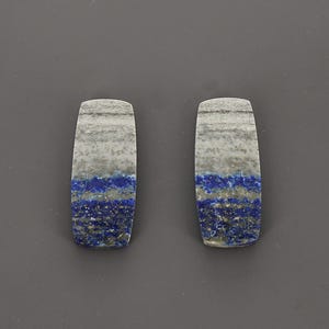 May include: Two rectangular lapis lazuli stones with rounded ends. The stones feature a mix of gray, white, and deep blue hues, with a layered appearance. The polished surfaces showcase the natural patterns and textures of the gemstone.
