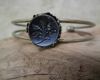 May include: A brass bangle bracelet with a dark blue button featuring a floral design with silver accents.