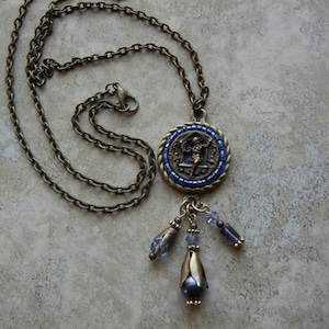 May include: A bronze chain necklace with a round pendant featuring a blue and gold mosaic design. The pendant has a small figure in the center and is accented with blue and purple crystal beads.