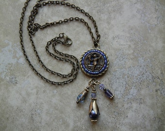 May include: A bronze chain necklace with a round pendant featuring a blue and gold mosaic design. The pendant has a small figure in the center and is accented with blue and purple crystal beads.