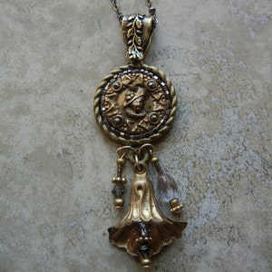 May include: A gold pendant necklace with a round, ornate charm featuring a detailed design. The charm is attached to a chain with a bell-shaped pendant and clear crystal beads.