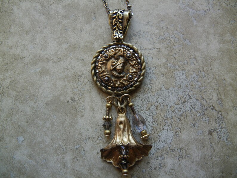 May include: A gold pendant necklace with a round, ornate charm featuring a detailed design. The charm is attached to a chain with a bell-shaped pendant and clear crystal beads.