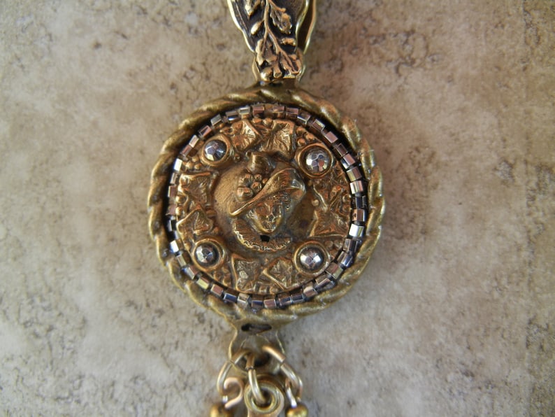 May include: A gold-toned pendant with a detailed design featuring a central circular motif with a raised, textured surface. The pendant is surrounded by a border of silver-toned beads.