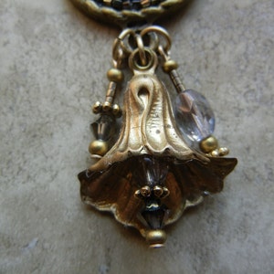 May include: A gold-tone pendant with a bell-shaped design and clear crystal accents. The pendant is adorned with small gold beads and hangs from a chain.
