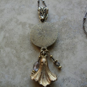 May include: A gold-toned necklace with a round pendant featuring a textured design. The pendant is attached to a chain with a small, ornate charm. The necklace also features a dangling charm shaped like a ghost.