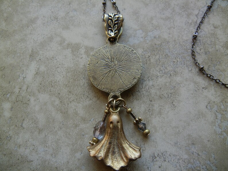 May include: A gold-toned necklace with a round pendant featuring a textured design. The pendant is attached to a chain with a small, ornate charm. The necklace also features a dangling charm shaped like a ghost.