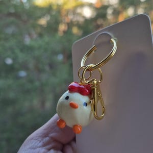 Puede incluir: Un llavero caprichoso con un pequeño pollito redondo con un lazo rojo, patas naranjas y un cierre y un llavero dorados. El pollito tiene ojos negros y un pequeño pico naranja. El fondo está borroso con tonos verdes y amarillos.