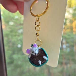 May include: A gold-colored keychain with a charm featuring a sleeping mouse. The mouse has white fur, pink ears, and is nestled in a brown pouch with a teal border. The keychain is attached to a white tag.