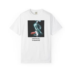 May include: White t-shirt featuring a square graphic of a human skeleton with a red brain, set against a dark background. The text "CRITICAL THINKER" is printed below the graphic in white. The shirt has a crew neck and short sleeves.