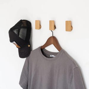 Könnte beinhalten: Drei Wandhaken aus Holz mit natürlicher Oberfläche sind an einer weißen Wand montiert. Eine schwarze Trucker-Mütze aus Mesh und ein graues T-Shirt an einem Holzbügel hängen an den Haken. Die Haken sind rechteckig mit einem abgerundeten Knauf.