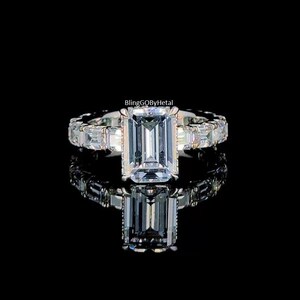 以下が含まれることがあります： 大きな長方形の透明な宝石を中央に配したシルバーリング。小さな長方形の宝石がバンドに沿ってセットされています。リングは黒い背景に表示され、下に反射が見えます。「BlingGQBbyHetal」というテキストが表示されます。
