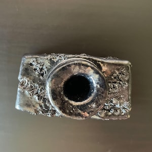 Puede incluir: Una escultura de cámara rectangular de color plateado con una lente circular grande. El cuerpo de la cámara tiene una superficie texturizada y rugosa. La lente es de color negro brillante.