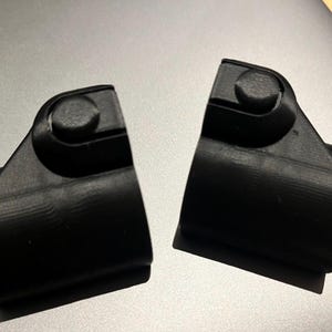May include: Two black, 3D-printed plastic components, possibly parts of a larger mechanism, are displayed. Each piece has a cylindrical base and a rounded top with a small, circular knob. The items are set against a gray background.
