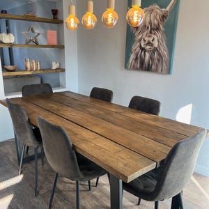 May include: A rustic wooden dining table with black metal legs, surrounded by six grey upholstered chairs. The table is set in a dining room with a painting of a Highland cow on the wall and a light fixture with four Edison bulbs hanging above.