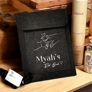 Puede incluir: Una funda de libro de fieltro negro con solapa. Presenta un dibujo lineal blanco de manos sosteniendo un anillo, con el texto "Myah's Red Book" y un corazón. También se ven una botella y un recipiente cilíndrico.
