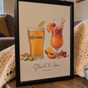Op de afbeelding: Ingelijste print met aquarel illustraties van een Corona bier en een cocktail met fruit garnituren. De print bevat de tekst "Corona" en de namen "Stewart & Jona EST. 2022" in een zwart frame.