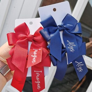 May include: Two ribbon hair bows, one red and one blue, each with a personalized name tag. The red bow has the name "Imogen" and the blue bow has the name "Alys" in white script. The bows are attached to white cards.