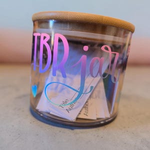 Puede incluir: Un tarro de cristal transparente con tapa de madera, etiquetado "TBR jar" en una fuente rosa y azul degradada. El tarro contiene trozos de papel, con una pequeña pegatina de corazón azul en el lateral. El tarro es para recomendaciones de libros.