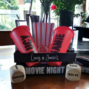 May include: A black wooden movie night box set with two red cups, a popcorn container, and dice. The cups have the names "Lucy" and "Jamie" printed on them. The dice have the words "Indian" and "Sci Fi" printed on them.