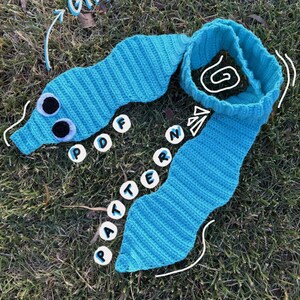 Puede incluir: Una bufanda de ganchillo turquesa con forma de gusano y grandes ojos saltones. La bufanda está sobre hierba y tiene las palabras "GIANT!!!" y "PDF PATTERN". Un accesorio único hecho a mano.