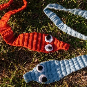 May include: Crocheted worms in red and light blue, with white and black eye details, are arranged on grass. The worms have a ribbed texture and long, slender bodies, ideal for a playful touch.