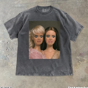 Puede incluir: Una camiseta gris carbón de manga corta con un estampado gráfico central de dos muñecas con expresiones de sorpresa. La muñeca de la izquierda tiene el pelo rubio y un top rosa, mientras que la de la derecha tiene el pelo castaño y un top dorado.
