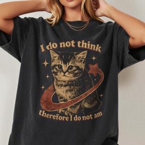Könnte beinhalten: Ein schwarzes Kurzarm-T-Shirt mit einer Grafik eines Kätzchens, das von einem Ring und Sternen umgeben ist. Der Text lautet, I do not think therefore I do not am, in einer Vintage-Schriftart. Das T-Shirt hat ein verwaschenes Aussehen.