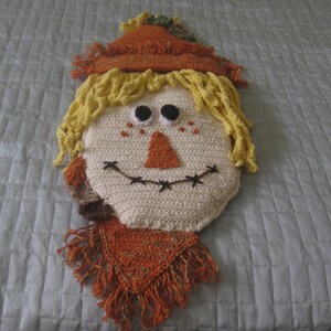 May include: A crocheted scarecrow pillow with a yellow straw hat, brown eyes, and a stitched smile. The pillow is made of beige, brown, and yellow yarn.