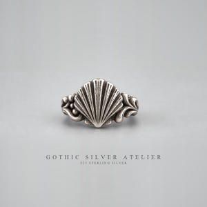 May include: A sterling silver ring featuring a central seashell motif with detailed vertical ridges, flanked by decorative scrollwork and small spheres on each side. The text GOTHIC SILVER ATELIER 925 STERLING SILVER is printed below the ring.