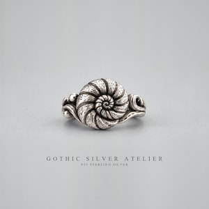 Könnte beinhalten: Ein Ring aus Sterlingsilber mit einem zentralen, von Ammonitenfossilien inspirierten Spiraldesign und strukturierten Details. Das Band ist an den Seiten mit kleinen kugelförmigen Akzenten verziert. Der Text GOTHIC SILVER ATELIER 925 STERLING SILVER ist sichtbar.