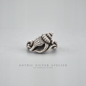 Può includere: Un anello in argento sterling caratterizzato da un design dettagliato a forma di conchiglia con scanalature strutturate e motivi a spirale. Il metallo presenta una finitura ossidata per evidenziare i dettagli intricati. Sotto l'anello, il testo recita GOTHIC SILVER ATELIER 925 STERLING SILVER.