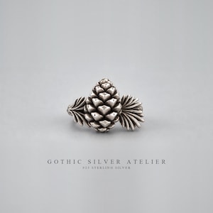 May include: A sterling silver ring featuring a central pinecone motif flanked by two stylized leaf-like structures. The metal has an oxidized finish to highlight the texture. Below the ring, the text reads GOTHIC SILVER ATELIER 925 STERLING SILVER.