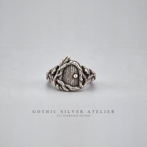 Puede incluir: Un anillo de plata de ley con un diseño de puerta arqueada central con detalles de veta de madera vertical y un pequeño pomo de cuenta redonda. La banda está esculpida con intrincados motivos de enredaderas y hojas. Texto: GOTHIC SILVER ATELIER 925 STERLING SILVER.