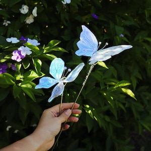 May include: Two delicate, light blue butterfly decorations on slender stems. The butterflies have translucent wings and are held against a backdrop of green foliage and sunlight. These decorative butterflies are ideal for crafts or home decor.