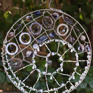 May include: A circular wall hanging featuring a silver wire frame in a spiral design. The structure is adorned with various dark grey and black marbled rings, polished stone beads, and small white pearls strung along the silver wire segments.