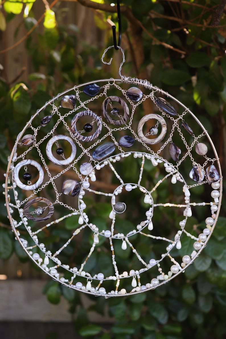 May include: A circular wall hanging featuring a silver wire frame in a spiral design. The structure is adorned with various dark grey and black marbled rings, polished stone beads, and small white pearls strung along the silver wire segments.