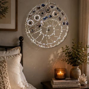 May include: A circular wall hanging featuring a silver wire frame adorned with dark blue, grey, and clear glass beads arranged in a spiral pattern. The piece is suspended from a black metal hook and incorporates various bead shapes and sizes.